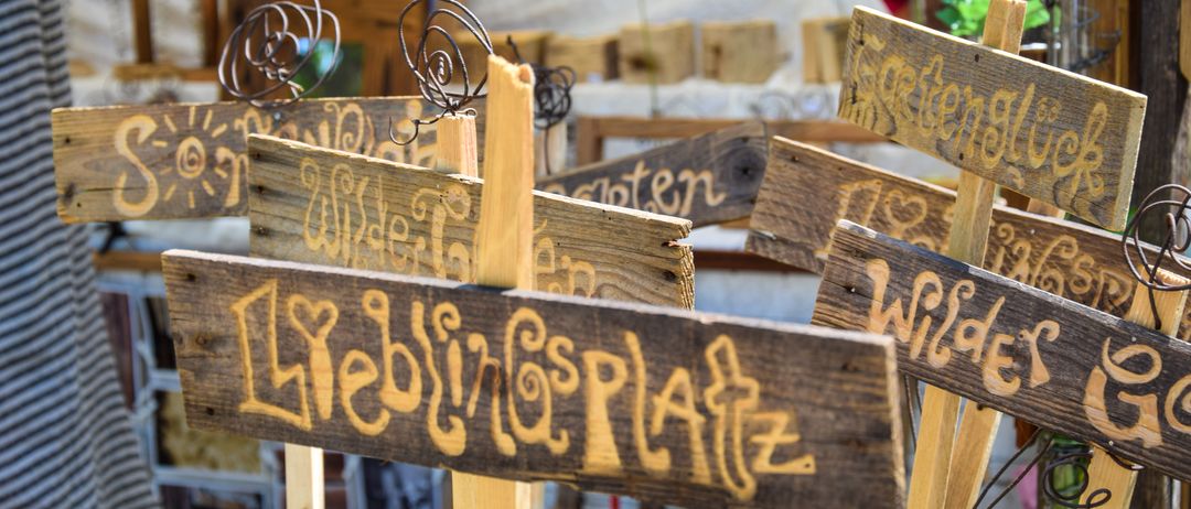 Eine Sammlung von Holzschildern und Handwerksarbeiten mit handgeschriebenem Text, auf einem Tisch ausgestellt. Eine Frau in einem rosa Shirt sitzt im Hintergrund, und ein Mann in einem blauen Shirt steht in der Nähe.