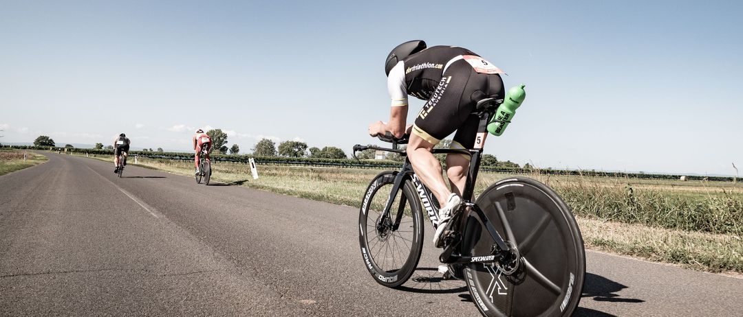 Ein Radfahrer mit schwarzem Helm fährt auf einer Straße mit einem Fahrrad, dahinter zwei weitere Radfahrer. Der Radfahrer hat eine grüne Wasserflasche am Fahrrad befestigt.