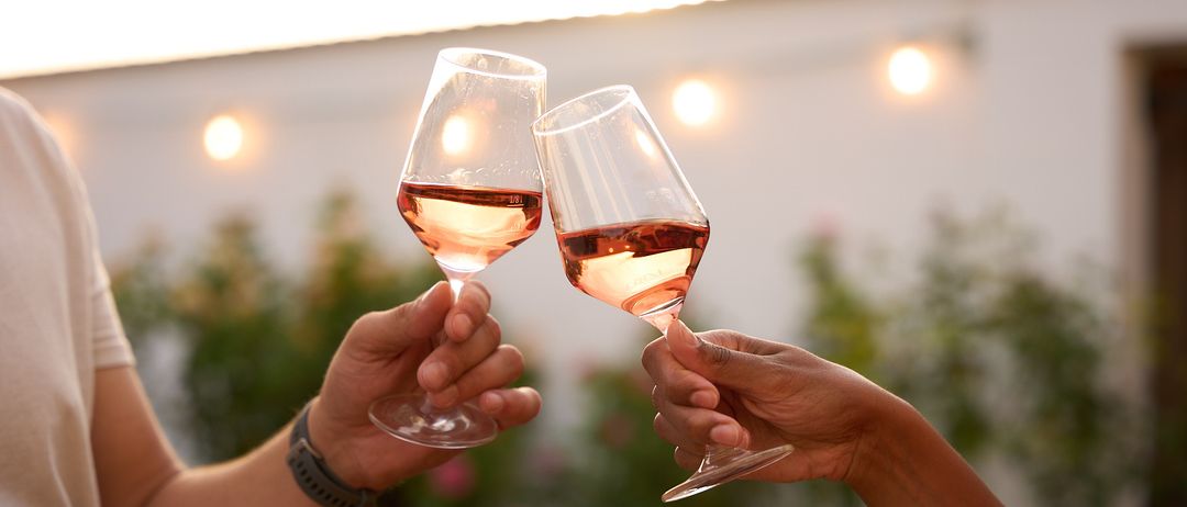 Zwei Personen stoßen mit Wein glasses an, halten sie nah aneinander und sind von einem Garten mit Lichterketten umgeben.
