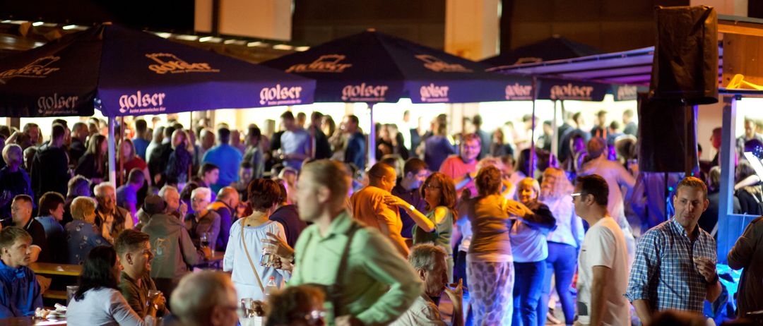 Eine große Gruppe von Menschen versammelt sich bei Nacht auf einem Außenbereich unter blauen Schirmen. Viele tanzen, einige stehen herum und einige sitzen. Die Atmosphäre ist lebhaft.