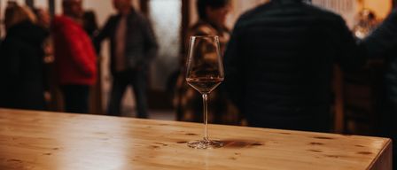 Ein mit rotem Wein gefülltes Weinglas steht auf einem Holztisch, im Hintergrund sind Menschen verschwommen.