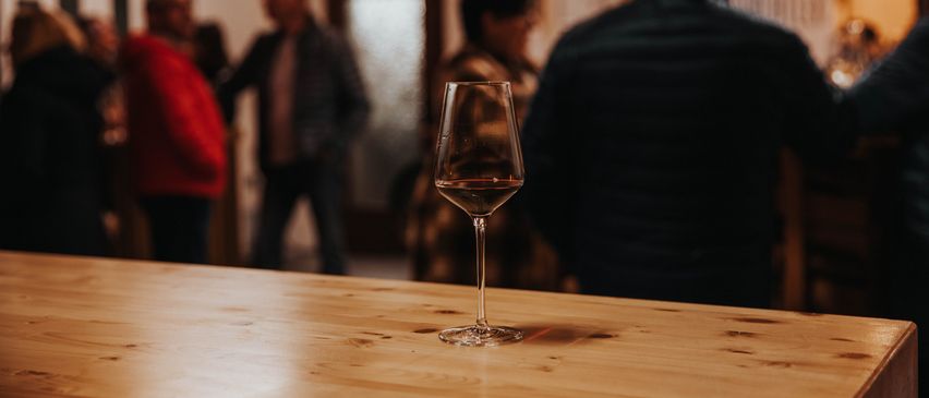 Ein mit rotem Wein gefülltes Weinglas steht auf einem Holztisch, mit verschwommenen Figuren von Menschen im Hintergrund.