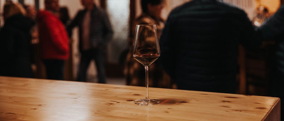Ein mit rotem Wein gefülltes Weinglas steht auf einem Holztisch, mit verschwommenen Figuren von Menschen im Hintergrund.