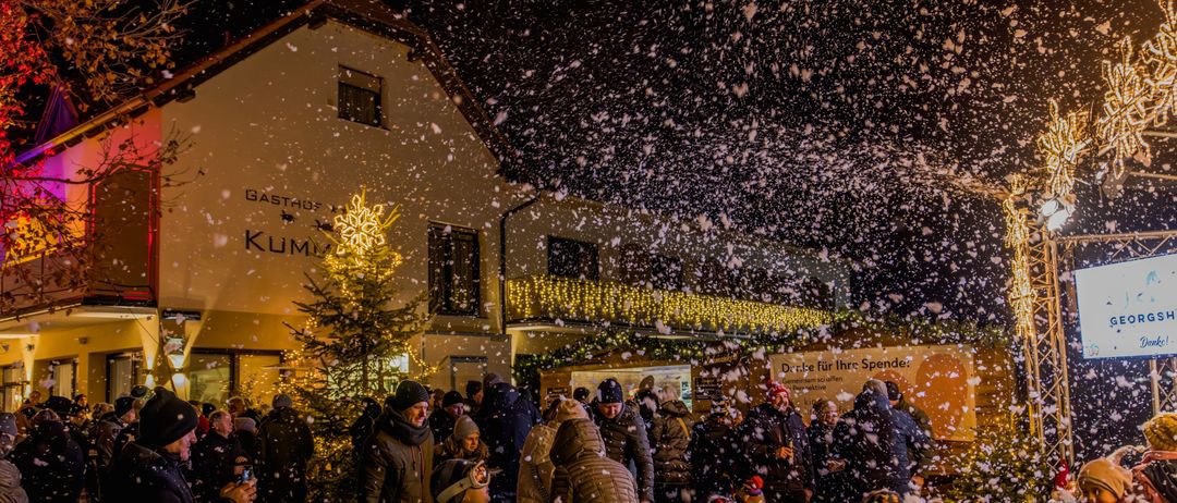 Ein festliches Treffen unter einem schneienden Himmel vor einem Gebäude, mit einem beleuchteten Weihnachtsbaum und einem Schild, das 'Getränke für Ihr Ausgeben' sagt.