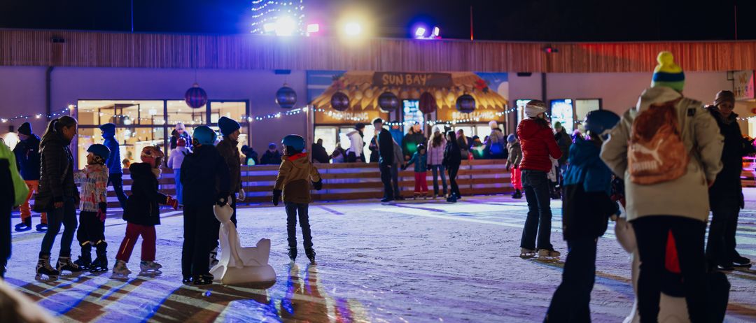 Eine Gruppe von Menschen eisläuft bei Nacht auf einer Eisbahn, einige stehen und andere rollen. Hinter ihnen befindet sich ein Gebäude mit einem Weihnachtsbaum und Lichtern.