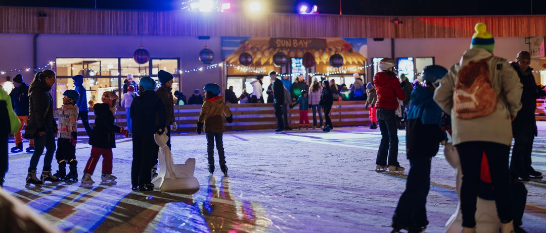 Viele Kinder Schlittschuh laufen auf einer Eisbahn im Freien bei Nacht. Sie tragen Winterkleidung. Ein Gebäude mit einem Weihnachtsbaum befindet sich hinter ihnen.