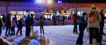 Viele Kinder Schlittschuh laufen auf einer Eisbahn im Freien bei Nacht. Sie tragen Winterkleidung. Ein Gebäude mit einem Weihnachtsbaum befindet sich hinter ihnen.