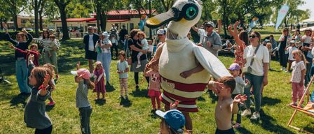 Ein großes Maskottchen in Form einer Ente unterhält eine Gruppe von Kindern und Erwachsenen in einem Park an einem sonnigen Tag.