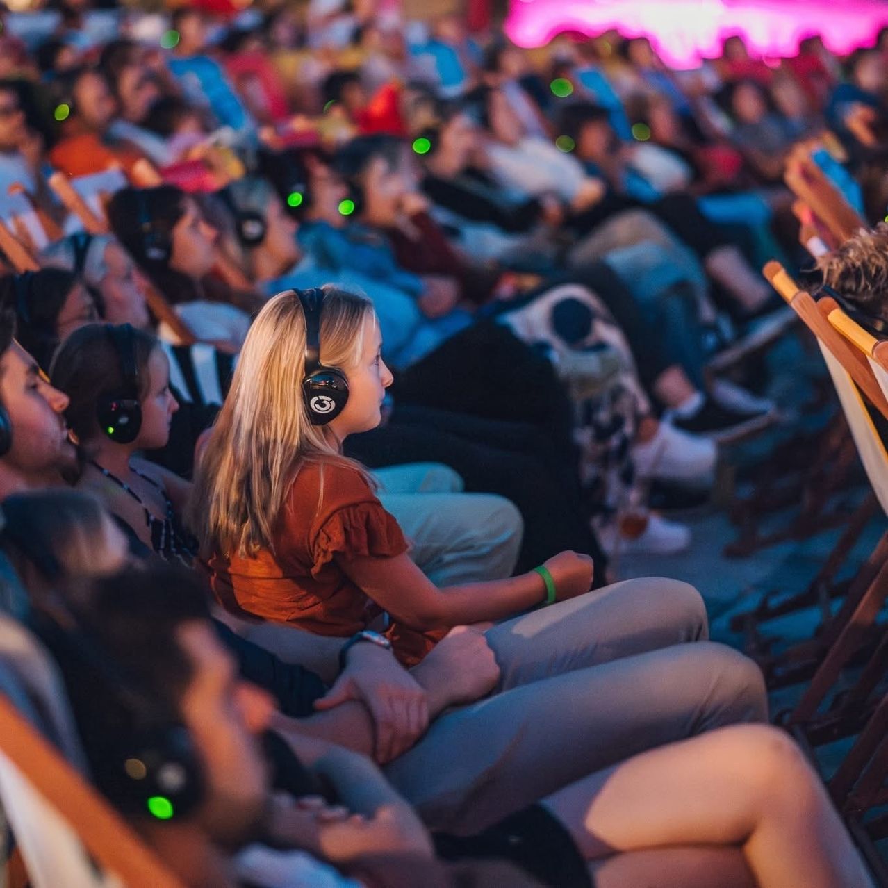 Eine Menschenmenge mit Kopfhörern sitzt in Reihen und sieht sich eine Präsentation oder einen Film an. Das Publikum scheint engagiert und konzentriert, einige nutzen ihre Handys.