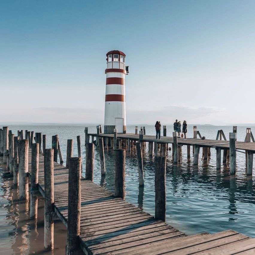 Ein Leuchtturm steht auf einem Pier, umgeben von einer Gruppe von Menschen. Der Pier erstreckt sich in ruhiges Wasser unter einem klaren blauen Himmel.