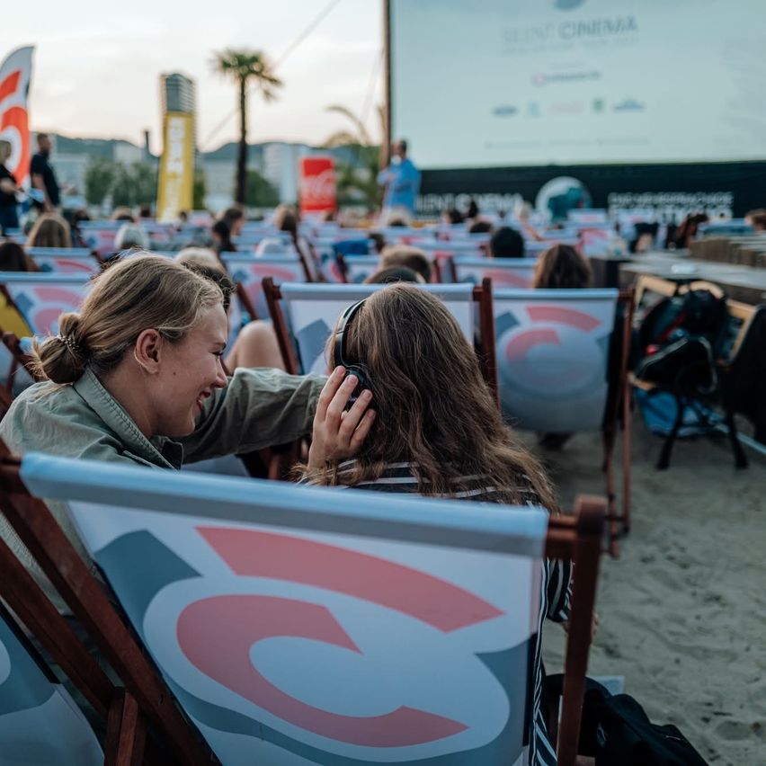 Zwei Frauen sitzen auf Strandstühlen, eine trägt Kopfhörer. Sie lachen, umgeben von anderen in einer Kinoumgebung. Im Hintergrund zeigt eine große Leinwand einen Film.