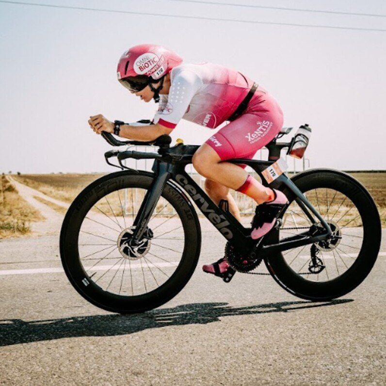 Ein Radfahrer fährt auf einer asphaltierten Straße und trägt ein pinkes Fahrradtrikot und einen Helm mit dem Wort 'BIOTIC'. Das Fahrrad ist mit 'Cervelo' beschriftet und mit einem Wasserflaschenhalter ausgestattet.