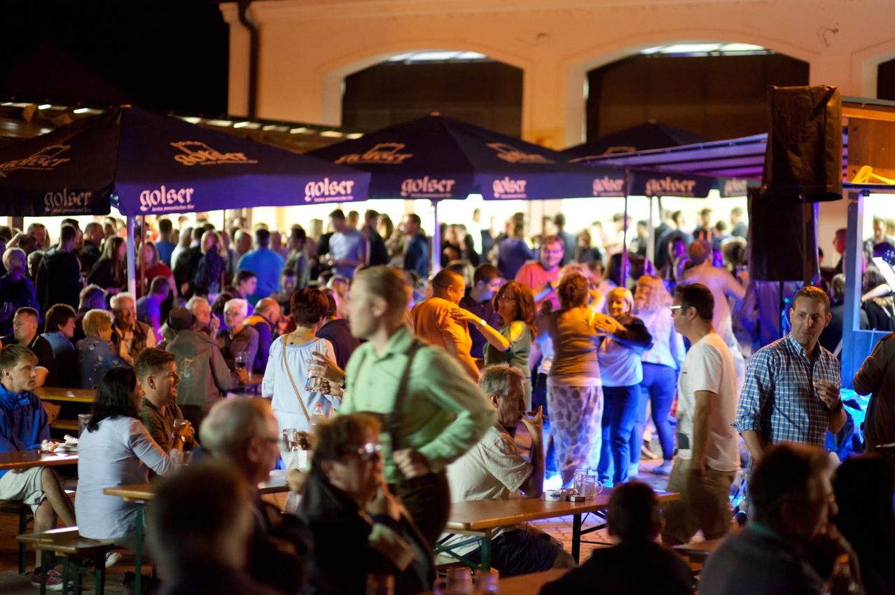 Ein überfüllter Außenbereich mit mehreren Schirmen, Menschen tanzen und plaudern, Tische und Stühle sind für Essen und Trinken aufgestellt, die Atmosphäre ist lebhaft und festlich.