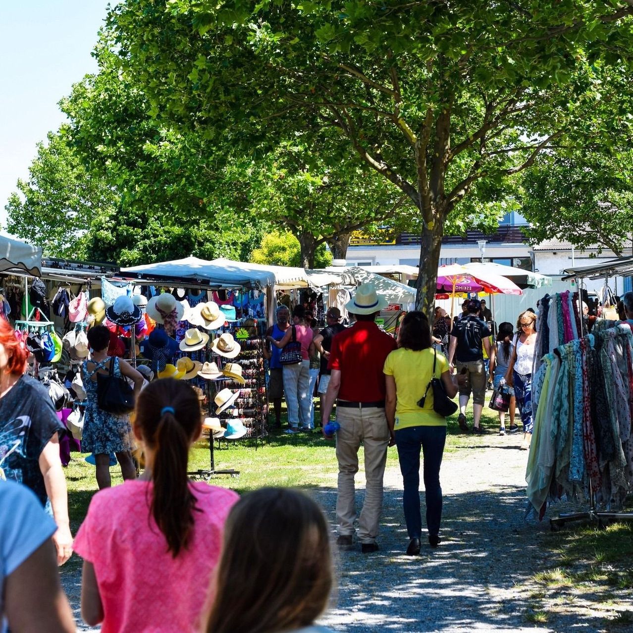 Ein Freiluftmarkt mit vielen Ständen, einkaufenden Menschen und unter Zelten ausgestellten Hüten. Bäume und Grün umgeben das Gebiet.