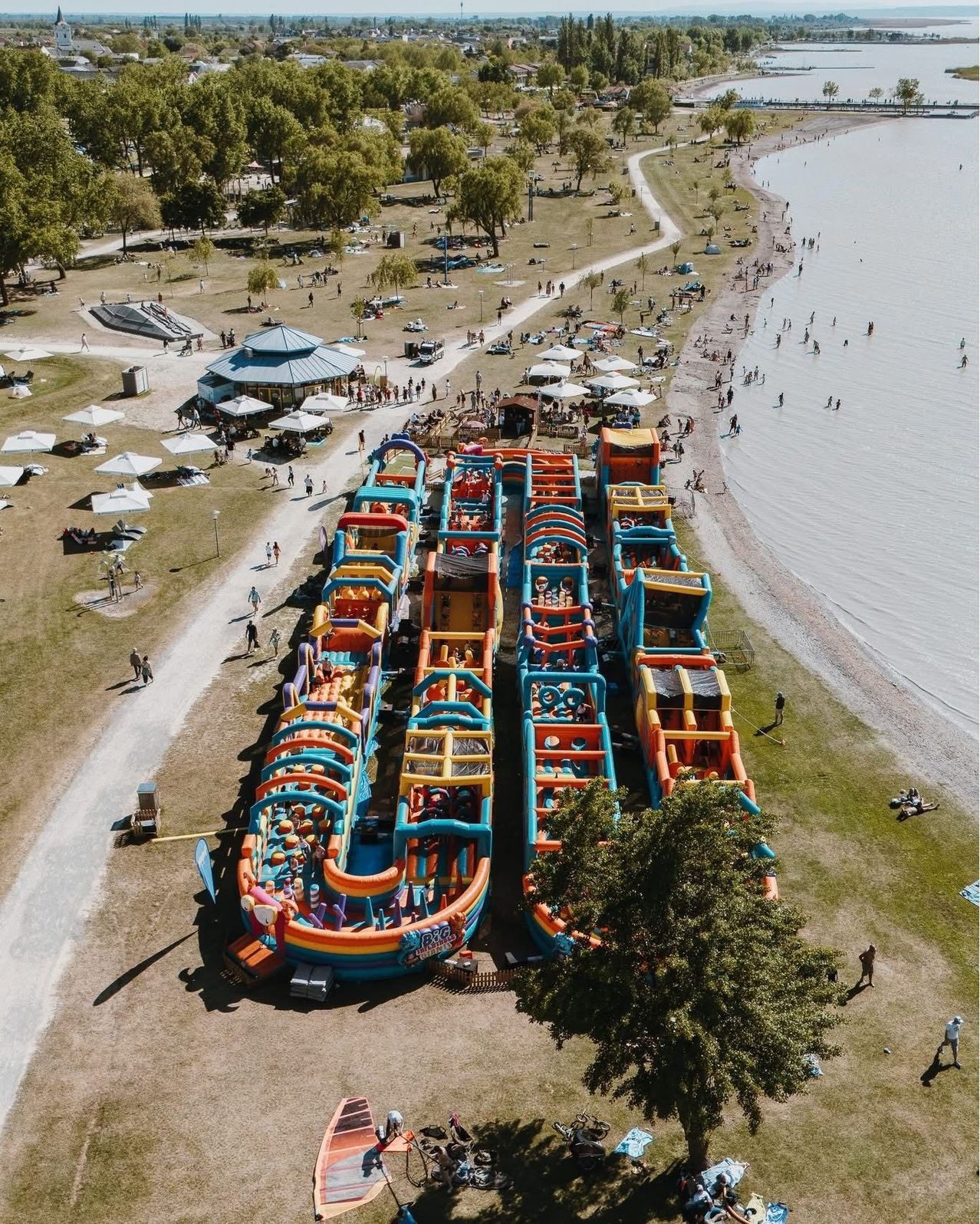 Ein Luftbild von einem Strand mit aufblasbaren Fahrattraktionen, Sonnenschirmen und Menschen, die am Ufer entlanggehen. Bäume und ein See sind im Hintergrund zu sehen.