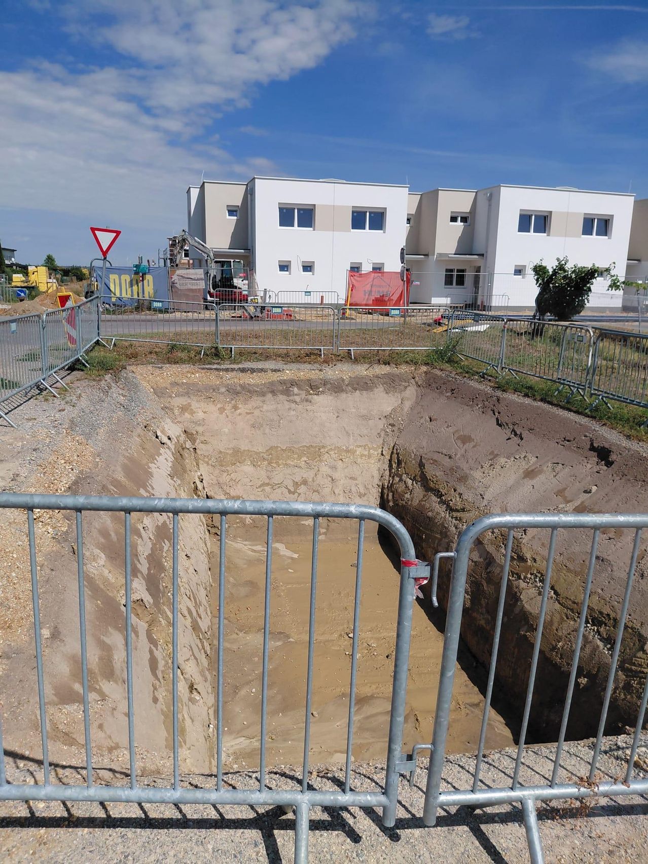 Eine Baustelle mit einem großen, eingezäunten Loch, einem weißen Gebäude im Hintergrund und Maschinen in der Nähe.
