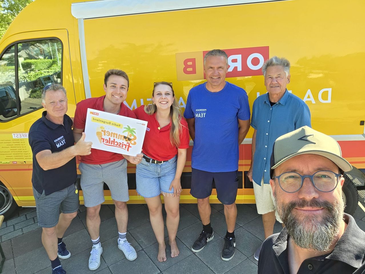 Fünf Personen posieren für ein Foto vor einem gelben Truck. Der Mann in der Mitte hält ein Schild mit 'Eisbecher' und einer lächelnden Frau neben ihm. Zwei Männer tragen blaue T-Shirts, und ein weiterer Mann mit einer Brille macht ein Selfie.
