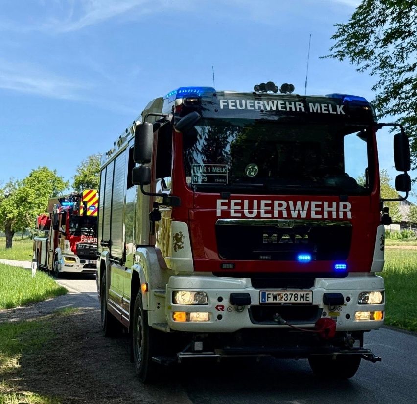 Ein roter Feuerwehrwagen fährt auf einer Straße mit einer grasbewachsenen Schulter. Ein zweiter Feuerwehrwagen folgt dahinter.