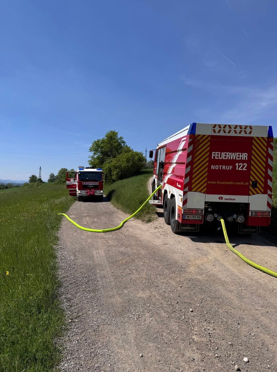 Ein Feuerwehrauto ist auf einem Feldweg geparkt, an dem ein gelber Schlauch angeschlossen ist. Ein weiteres Feuerwehrauto ist dahinter geparkt. Ein Grasfeld mit einigen Bäumen befindet sich auf der linken Seite.