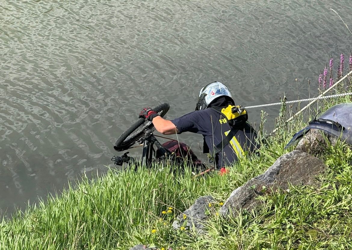 Eine Person in einem Sicherheitshelm und Handschuhen hält ein Fahrrad am Rad, teilweise im Wasser eingetaucht, mit einem Grasufer in der Nähe.