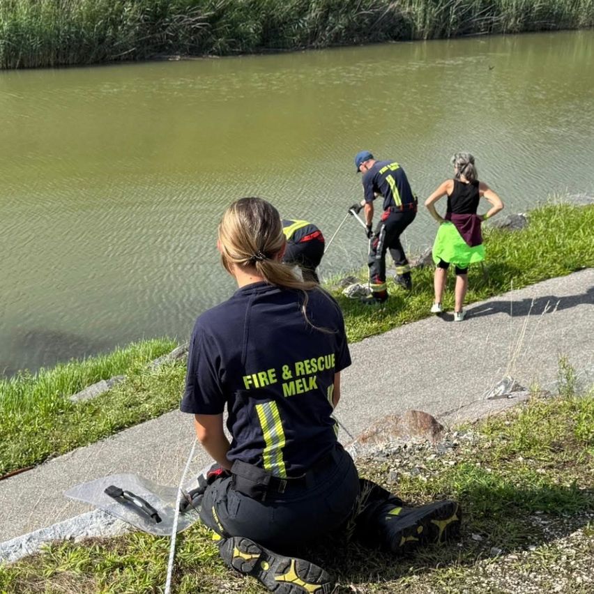 Drei Personen, zwei in Feuerwehruniformen, befinden sich in der Nähe eines Gewässers. Eine Person sitzt auf dem Gras, und zwei andere scheinen eine Inspektion durchzuführen.