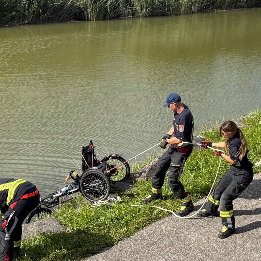 Drei Feuerwehrleute ziehen einen Rollstuhl aus dem Wasser. Einer steht am Ufer und zwei auf der Straße. Der Fluss ist ruhig.