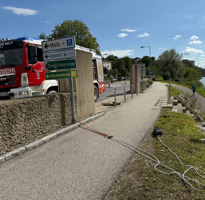Ein Feuerwehrwagen ist am Straßenrand geparkt, mit Kabeln und Stangen in der Nähe des Bürgersteigs. Straßenschilder zeigen die Richtungen nach Melk und zum Bahnhof an.