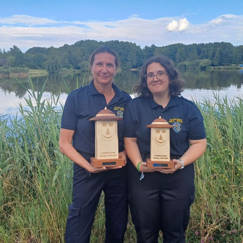 Zwei Frauen stehen vor einem See, halten Trophäen und lächeln für ein Foto. Sie tragen passende Uniformen mit Abzeichen.