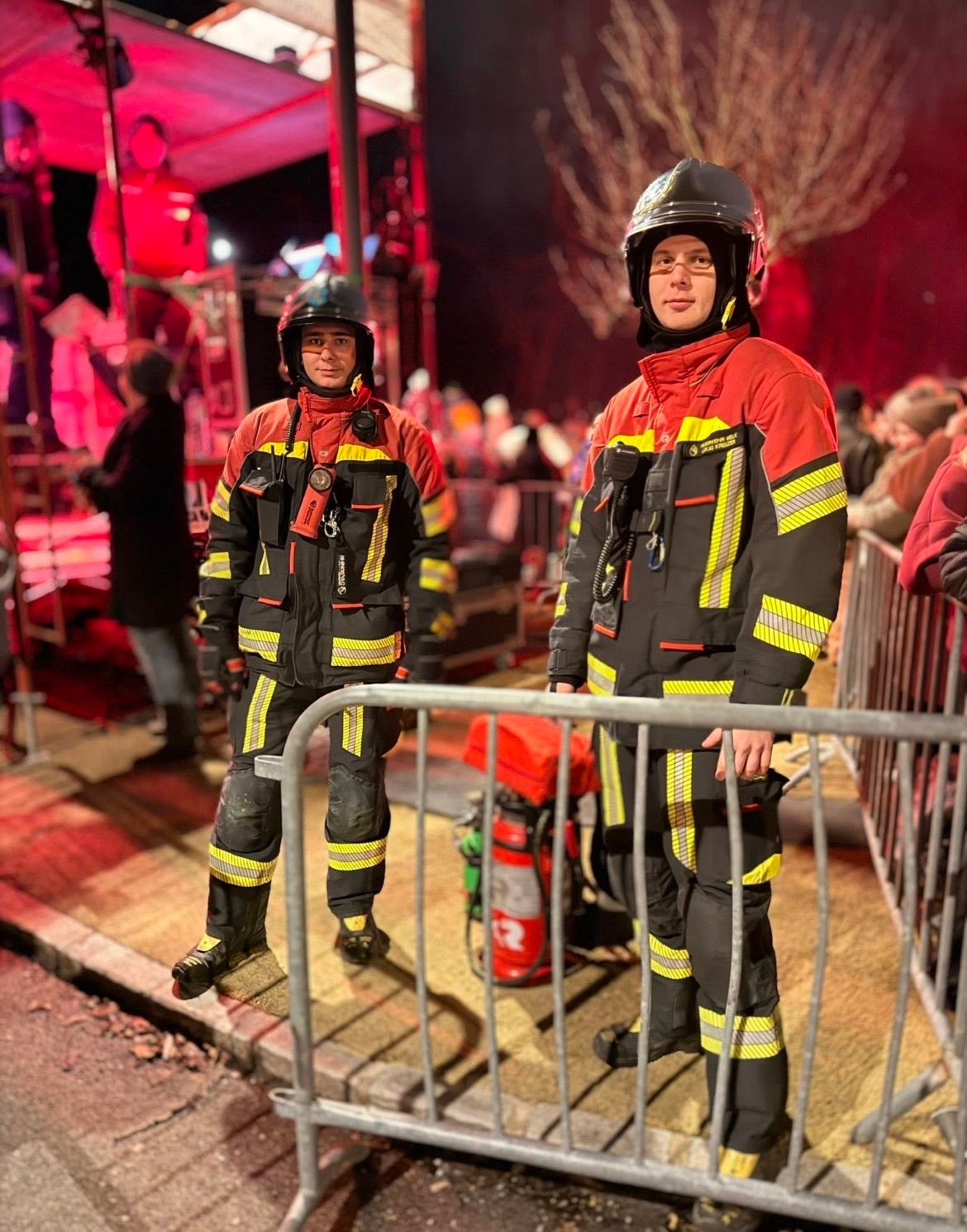 Zwei Feuerwehrleute stehen nebeneinander in roten und schwarzen Uniformen mit gelben Streifen. Sie stehen in der Nähe einer Metallbarriere mit einem Feuerlöscher dahinter. Eine Menge beobachtet sie auf der Straße.