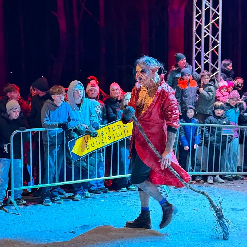 Ein als Hexe verkleideter Schauspieler führt vor einer Gruppe von Kindern auf der Bühne auf, hält einen Besen und ein gelbes Schild in der Hand.