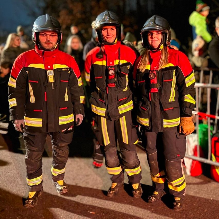Drei Feuerwehrleute in roten und schwarzen Uniformen mit gelben Streifen stehen zusammen auf einer Straße bei Nacht, mit einer Menschenmenge im Hintergrund.