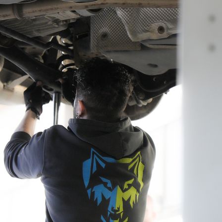 Eine Person arbeitet an der Unterseite eines Autos und repariert etwas mit einem Werkzeug. Die Person trägt eine blaue Kapuzenjacke mit Wolfsdesign.