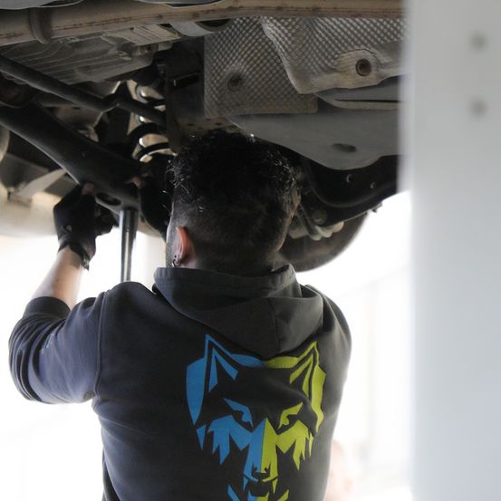 Eine Person arbeitet an der Unterseite eines Autos und repariert etwas mit einem Werkzeug. Die Person trägt eine blaue Kapuzenjacke mit Wolfsdesign.