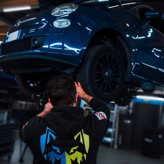 Ein Mann in einer schwarzen Kapuzenjacke arbeitet an einem blauen Auto mit Logo, wahrscheinlich in einer Garage. Das Auto ist in der Luft gehoben.