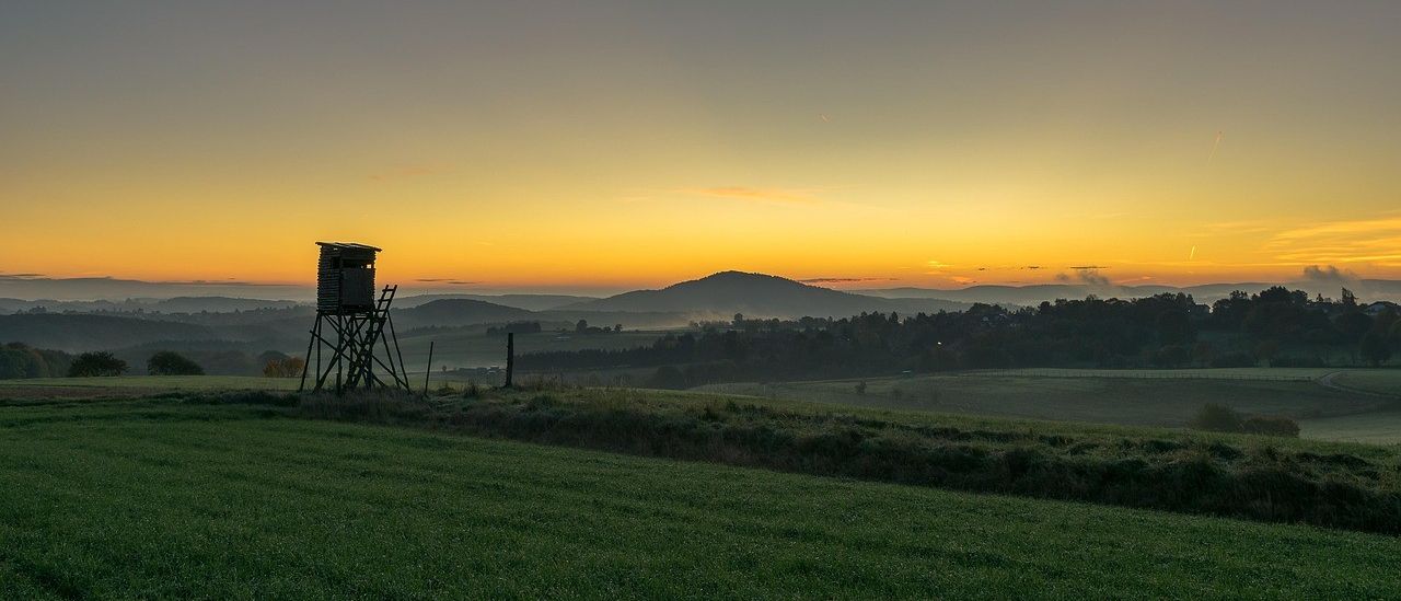 Eine ruhige Landschaft bei Sonnenuntergang mit einem Aussichtsturm in einem grünen Feld, Bergen in der Ferne und einem klaren Himmel.