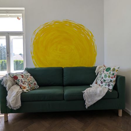 Ein Wohnzimmer mit einem grünen Sofa, Kissen und einer Sonnen-Wandkunst. Der Raum hat einen Holzboden und ein Fenster mit einem Schmetterling.
