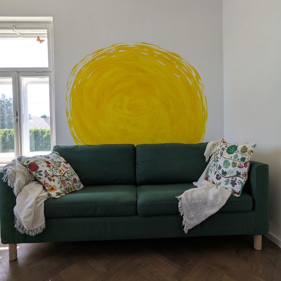 Ein Wohnzimmer mit einem grünen Sofa, Kissen und einer Sonnen-Wandkunst. Der Raum hat einen Holzboden und ein Fenster mit einem Schmetterling.