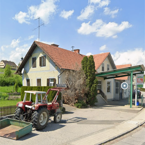 Ein roter Traktor steht vor einem Haus mit rotem Dach. Eine grüne Überdachung ist am Haus befestigt. Eine Treppe führt zum Eingang des Hauses. Ein Auto ist in der Ferne geparkt.