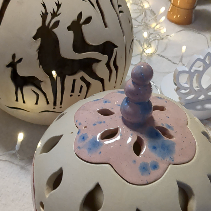 Zwei Keramikgläser sind ausgestellt. Eines hat eine Hirschsilhouette, das andere ein blaues und rosa Blumendesign mit einem lila Deckel.