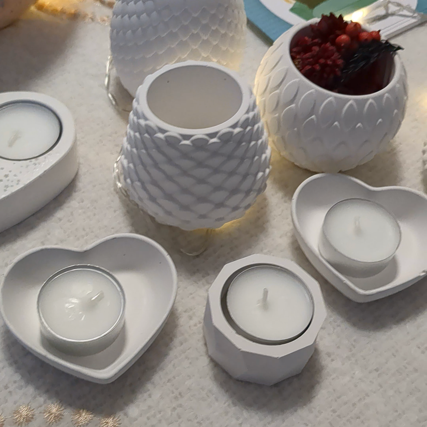 Mehrere weiße Keramik-Kerzenhalter auf einem Tisch, darunter herzförmige und sechseckige Designs, einige mit brennenden Kerzen. Ein weißer ananasförmiger Halter mit Beeren ist ebenfalls vorhanden.
