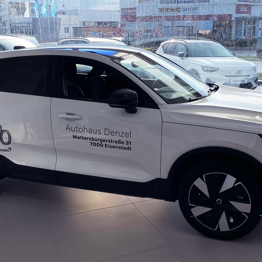 Ein weißer Pkw mit 'Autohaus Denzel' und seiner Adresse an der Seite steht in einem Ausstellungsraum mit anderen Autos und einem Gebäude im Hintergrund.