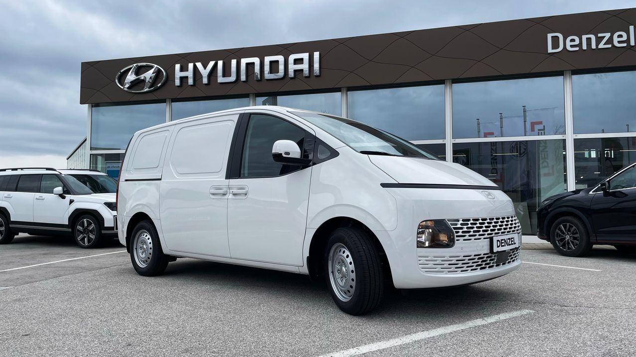 Ein weißer Hyundai-Transporter steht vor einem Hyundai-Händler. Der Van ist auf einem gepflasterten Parkplatz geparkt, über dem ein Hyundai-Schild angebracht ist.