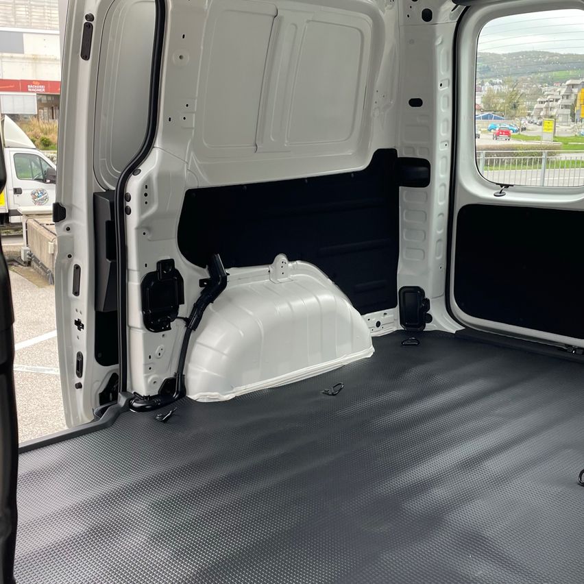 Innenansicht eines weißen Lieferwagens mit geöffneten Türen. Der Laderaum hat einen schwarzen Boden und eine weiße Aufbewahrungsbox. Die Seitenwände sind schwarz und das Heckfenster zeigt eine Stadtansicht mit Gebäuden und einer Straße.