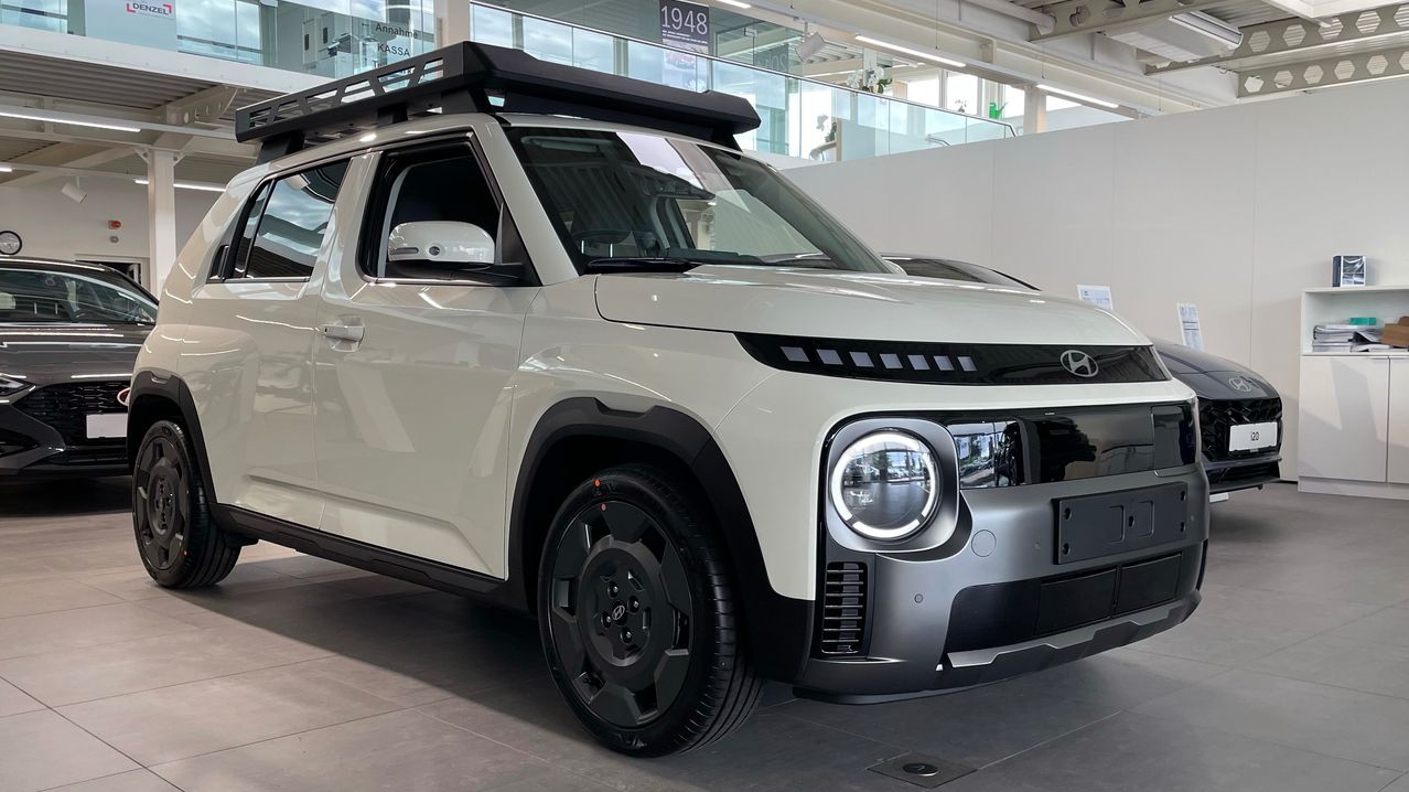 Ein weißer Hyundai SUV mit Dachgepäckträger und schwarzem Stoßfänger wird in einer Ausstellung gezeigt. Das Auto verfügt über moderne LED-Scheinwerfer und einen geräumigen Innenraum.