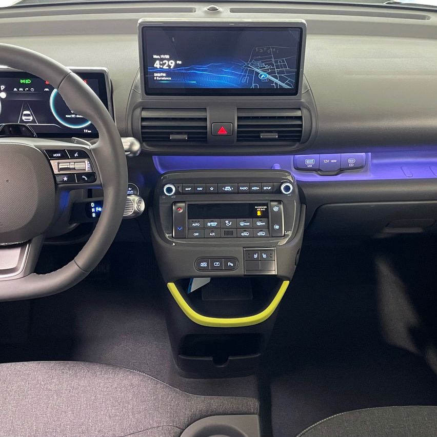 Der Innenraum eines modernen Autos zeigt ein Armaturenbrett mit einem Touchscreen, der die Zeit als 4:29 anzeigt. Das Lenkrad befindet sich auf der linken Seite, und das Armaturenbrett hat Steuerknöpfe und ein digitales Display. Der Innenraum des Autos ist dunkel mit grauen Sitzen und Akzenten.