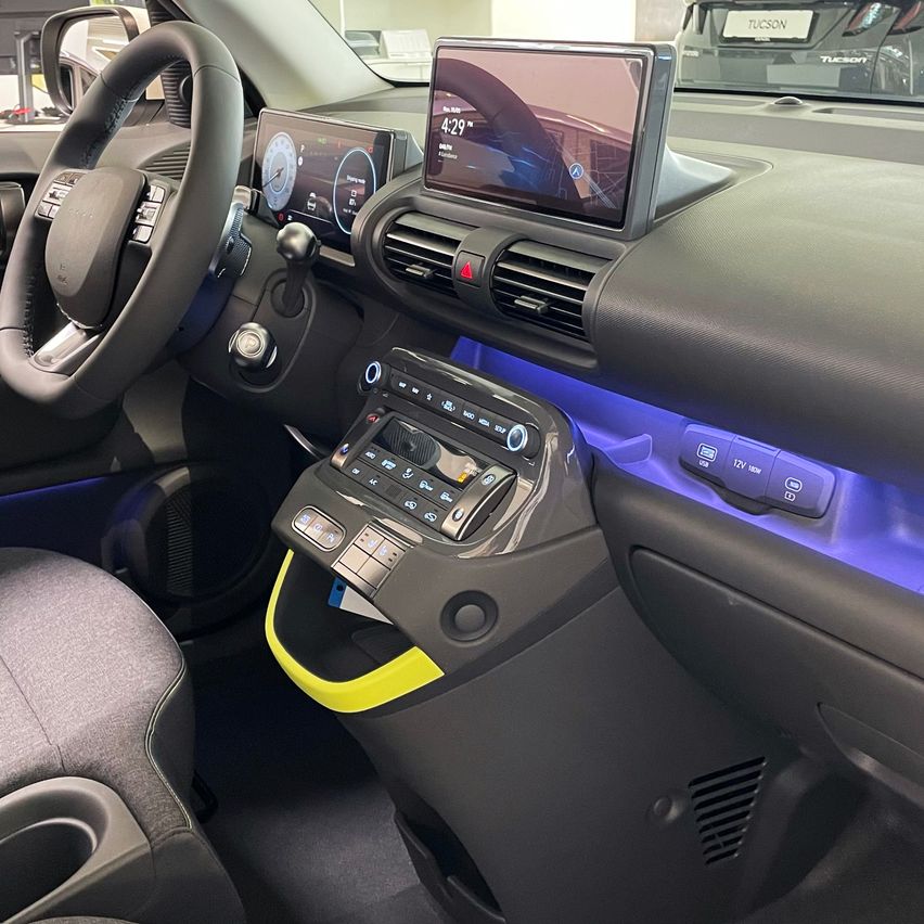 Innenraum eines modernen Autos mit einem Armaturenbrett, das einen großen zentralen Touchscreen, einen kleineren Bildschirm am Lenkrad und verschiedene Bedienknöpfe aufweist. Das Armaturenbrett hat blaue Akzentbeleuchtung.