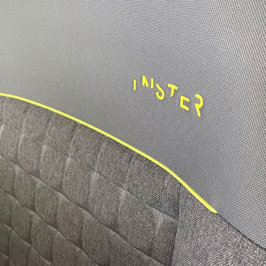 Eine Nahaufnahme eines grauen Stoffsitzes mit einer gelben Linie am Rand. Das Wort 'INISTER' ist in Gelb auf dem grauen Stoff geschrieben.