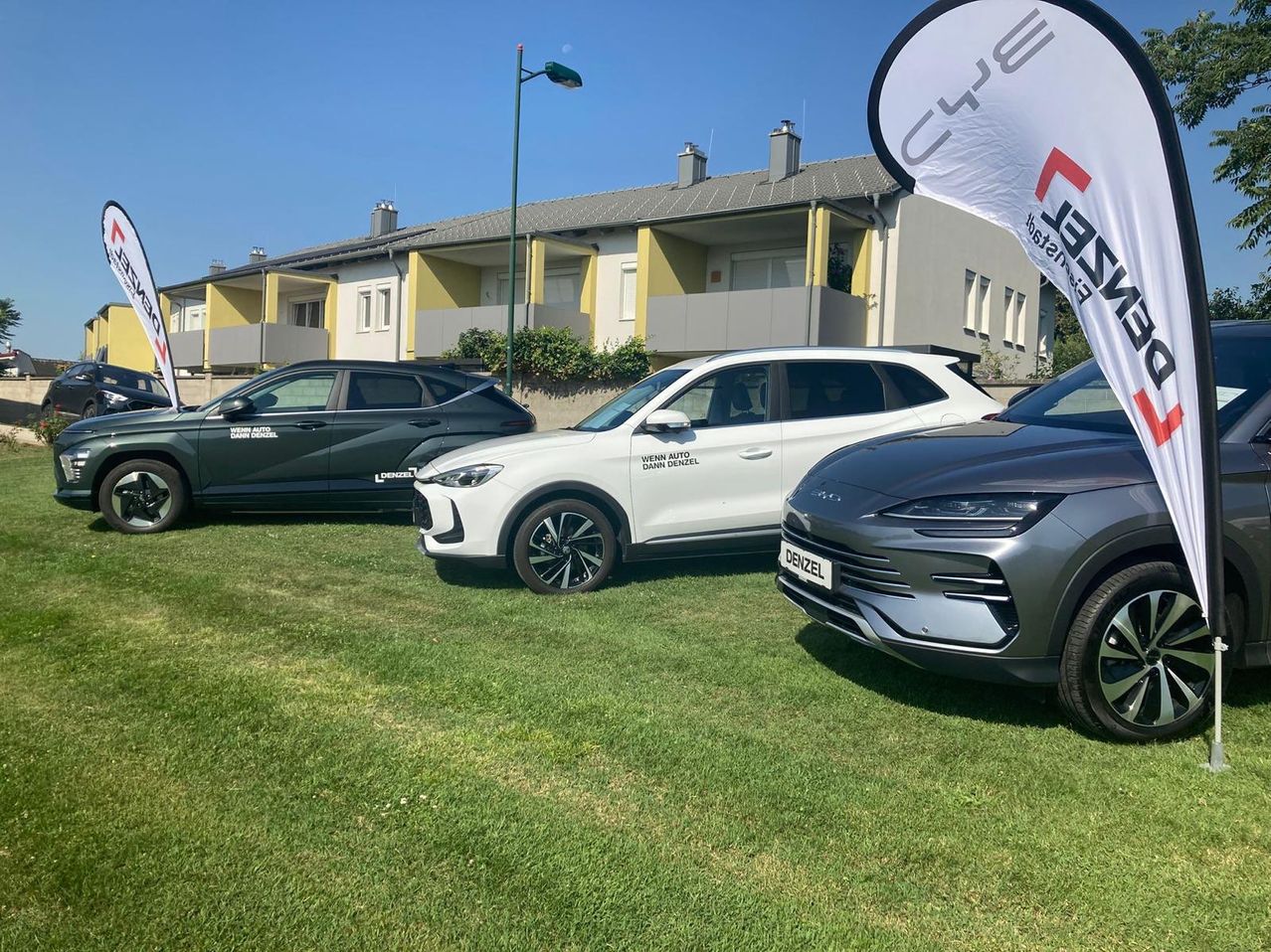 Drei Autos sind auf einem Rasen vor einem Gebäude geparkt. Ein weißes Banner mit Text steht vor einem grauen Auto.