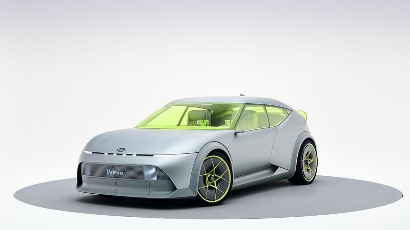 Ein futuristisches Konzeptauto mit schlankem Design, grauer Karosserie, neongrünen Akzenten und Speichenrädern wird auf einer grauen Plattform präsentiert.