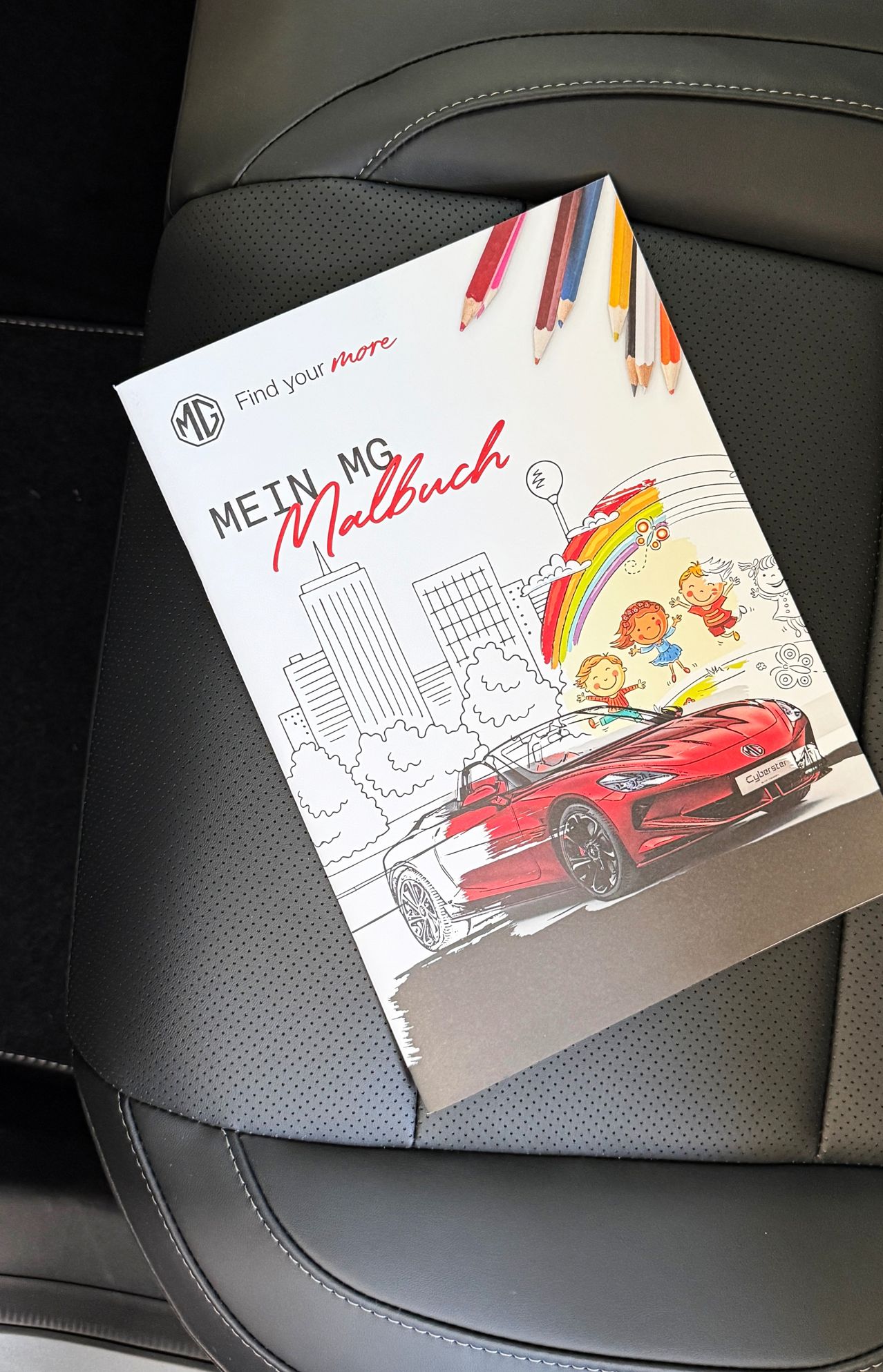 Ein Malbuch mit dem Titel 'Mein MG Malbuch', das einen roten Sportwagen und spielende Kinder auf dem Cover zeigt, auf einem schwarzen Autositz mit farbigen Bleistiften in der Nähe.